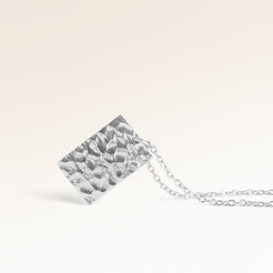 Närbild av minimalistiskt fyrkantigt berlock i silver med vågig yta och plats för gravyr baktill
