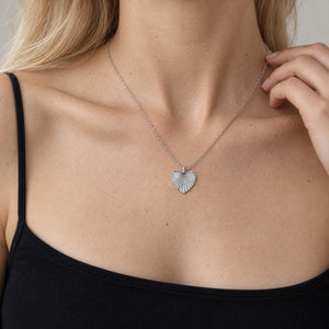 Modell i svart linne med smala axelband som rör vid kedjan på sitt hjärtformade silverhalsband med räfflat solstrålemönster och plats för personlig gravyr av text, koordinater eller handstil på baksidan